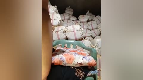 Video of a small, sparsely furnished room with a single green plastic chair, an orange and white floral pillow, and stacks of white and pink burlap sacks filled with goods, likely aid supplies.