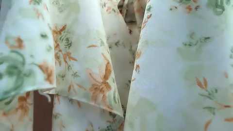 A close-up video of a woman's face partially obscured by floral-patterned fabric. The fabric is white with green and orange flowers, creating a soft, textured background. The woman's skin tone is light, and her eyes are visible, suggesting a serene expression.