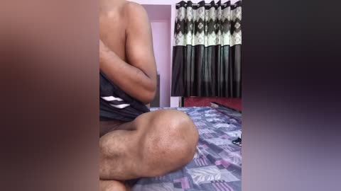 A video of a muscular, light-skinned man's upper body, partially dressed in a black shirt, sitting on a bed with patterned bedding. The background features curtains and a red wall.