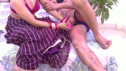 A video of an Indian woman in a maroon sari with white stripes, adorned with bangles, holding a man's penis, while he sits naked on a patterned blanket.