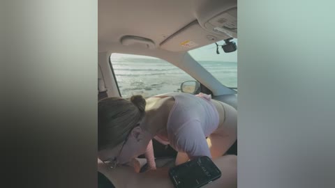 Video of a woman with light skin and brown hair in a ponytail, wearing a white tank top and white panties, leaning over a man's lap in a car. The man has a tattoo on his arm.