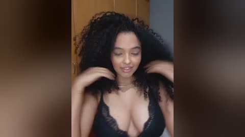 Video of a young, light-skinned Black woman with voluminous, curly hair, wearing a black lace bralette. She is adjusting her hair in a bedroom with wooden wardrobes in the background.