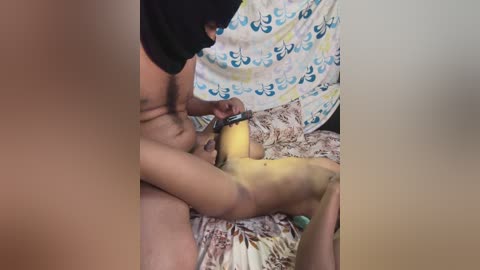 Media: A video showing a man with dark skin, wearing a black t-shirt, holding a yellow vibrator to a woman's vagina. She has light skin, and they are on a bed with a colorful, patterned sheet.