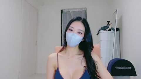 Video of an East Asian woman with long black hair, wearing a blue tank top, blue face mask, and seated in a room with a white door, laundry basket, and a black hat.