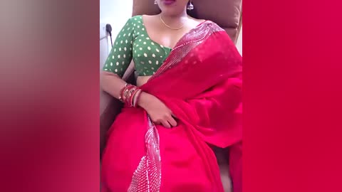 Video of a South Asian woman in a green polka-dotted blouse, red saree, gold necklace, and bangles, seated on a brown chair, partially obscured by a red curtain.