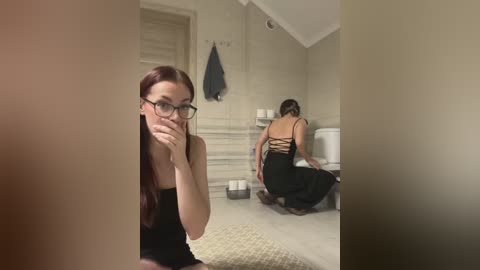 Video of a young woman with long red hair, wearing black glasses and a black dress, squatting in a beige-tiled bathroom, reflecting in a mirror.