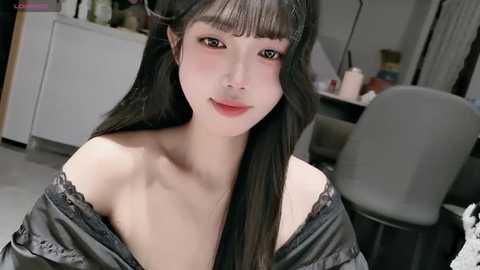 Video of an Asian woman with long black hair, wearing a black off-shoulder dress, sitting in a modern office with a desk and chair in the background.