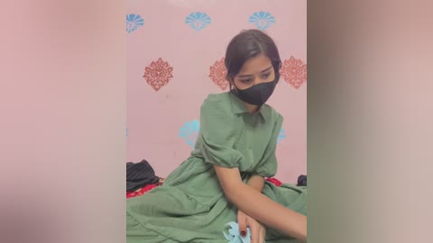 Video of a young woman with medium skin tone and dark hair, wearing a green dress, black mask, and blue headphones, seated on a bed against a pink wall adorned with blue and orange flower decals.