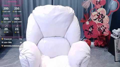 A video of a white, plush, armchair in a room filled with pink heart decorations and a blue curtain backdrop. Social media icons and a live stream overlay are visible.