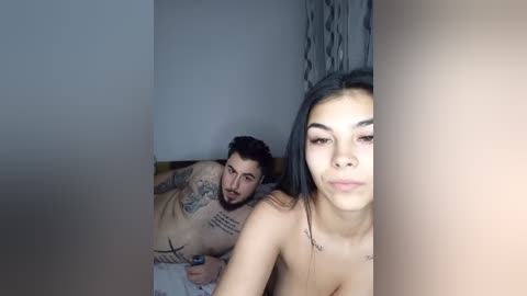 Video of a shirtless man with tattoos, lying on a bed in a dimly lit room, while a topless woman with long black hair, wearing makeup, stands partially hidden by a pillar.