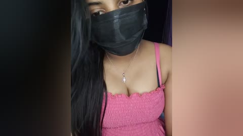 Video of a woman with long black hair, wearing a pink spaghetti-strap top, black mask, and a silver necklace. Her face is partially obscured by the mask.