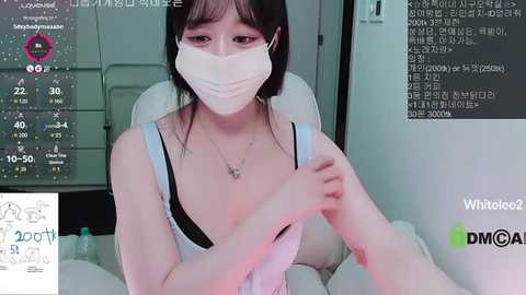 Video of an East Asian woman with pale skin, dark hair, and a mask, sitting on a white chair, holding a white device. Background shows a live stream with digital text and a logo.