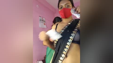 Video of a woman in a black saree with floral designs, wearing a red mask and drinking from a bottle. Pink walls, posters, and a green chair in the background.