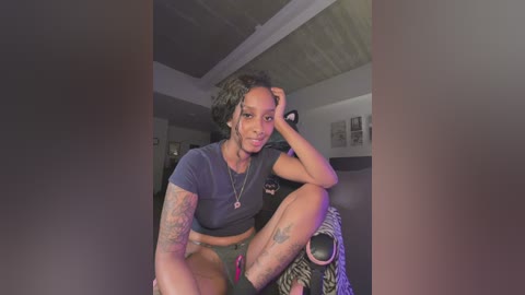 Video of a young, dark-skinned, tattooed woman with short, curly hair, wearing a black crop top and green shorts, sitting in a dimly lit room with exposed wooden ceiling.