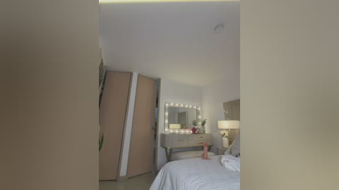 Video of a modern, minimalist bedroom with beige walls, white bedding, a wooden dresser with a mirror, and a lamp on the nightstand. The room features a large wardrobe with a partially open door.