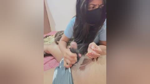 Video of an Asian woman with long black hair, wearing a mask, lying on a bed with a pink sheet, using a blue cloth to clean her vagina.
