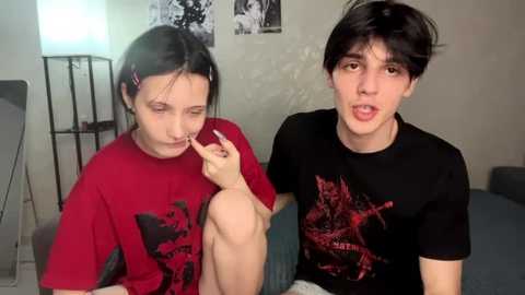 Video of two young people with pale skin, one with black hair wearing a red shirt, the other with brown hair in a black shirt, both looking solemnly at a cigarette.