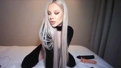 Video of a slender, platinum-blonde woman with long hair, wearing black lingerie, sitting seductively on a bed, eyes closed, surrounded by a beige wall and grey curtains.