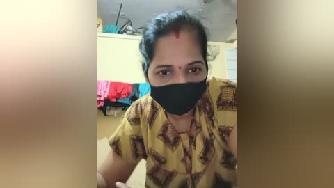 Video of a South Asian woman with medium skin tone, wearing a black face mask and yellow-patterned shirt, seated in a cluttered, dimly lit room with colorful clothes hanging on a rack in the background.