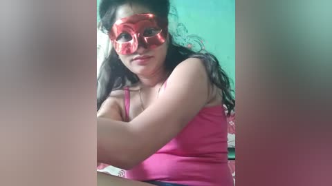 Video of a light-skinned woman with long black hair wearing pink glasses and a pink tank top, seated indoors with a teal wall in the background.