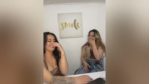 Video of two young women, one with a tattooed arm, sitting on a white couch. One woman is laughing, the other covering her face. A framed \"smile\" sign hangs on the white wall.