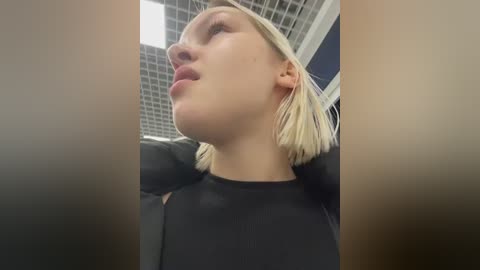 Media: Video of a young woman with fair skin, blonde bob haircut, and a black top, looking upward in a tiled ceiling room.