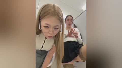 Media: Video of two young women in a bathroom; one with long blonde hair, glasses, and a white blouse, the other with dark hair, wearing a white top and black skirt, both appearing shocked.