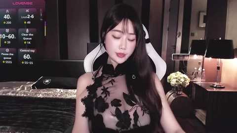 Video of an Asian woman with long black hair, wearing a black lace top, seated in a dimly lit room with gaming equipment and flowers.