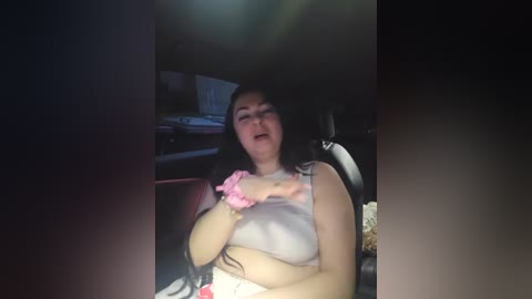 Video of a middle-aged woman with long dark hair, wearing a pink scrunchie and light gray tank top, seated in a dimly lit car, mouth open, possibly in mid-speech.