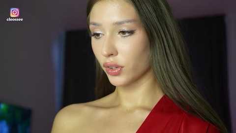 Video of an Asian woman with long, straight brown hair, wearing a red off-shoulder dress, looking contemplative. Background is blurred with dark tones, showcasing her light skin and subtle makeup.