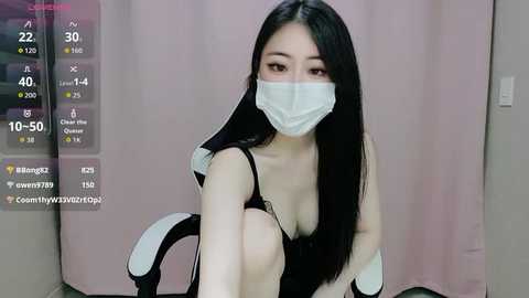 Video of an Asian woman with long black hair, wearing a black tank top, white mask, and gloves, sitting on a chair in a room with a pink curtain background.