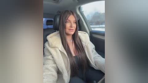 A video of a woman with long, straight brown hair, wearing a white fur coat and black top, seated in the driver's seat of a car.