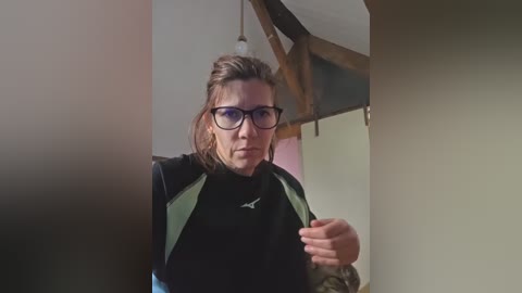 Video of a Caucasian man with a beard and glasses, wearing a black and green jacket, standing indoors with wooden beams and a light bulb visible in the background.