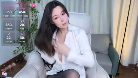 A young Asian woman with long black hair, fair skin, and medium breasts, wearing a white blouse unbuttoned to reveal black lingerie, sits in a modern living room.