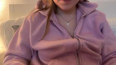 Video of a young woman with light skin and long red hair, wearing a lavender hoodie, a heart necklace, and sitting on a beige couch in a dimly lit room.