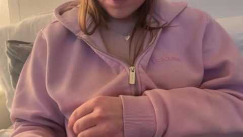 A video of a light-skinned woman with long brown hair, wearing a pastel pink hoodie, sitting on a beige couch. The background is indistinct, but the image focuses on her crossed arms and slight smile.