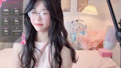 Video of a young East Asian woman with long black hair, glasses, and a white sleeveless top, smiling in a pastel-colored bedroom with a plush bear and digital clock.