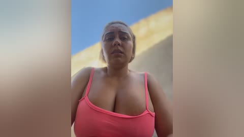 Video of a plus-size, light-skinned woman with blonde hair in a pink spaghetti-strap tank top, standing in front of a beige building with a blue sky background.