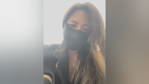 A video of an Asian woman with long, straight, brown hair, wearing a black face mask and a dark jacket, standing in a dimly lit, indoor hallway with blurred, white walls.