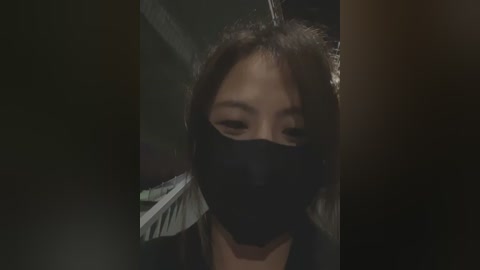 Video of a person with medium-length brown hair, wearing a black face mask, standing in a dimly lit room with dark walls and a blurred background.