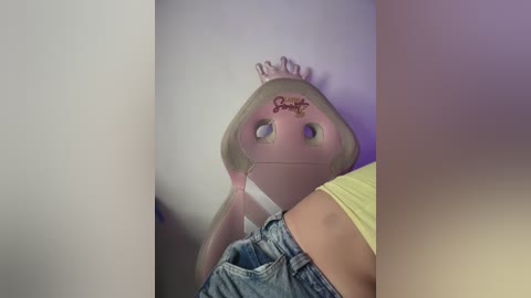 Video of a young girl wearing a pink, heart-shaped mask with \"Bonnie\" written in red cursive, light blue denim shorts, and a yellow shirt. The background is a soft purple wall, creating a playful and whimsical atmosphere.