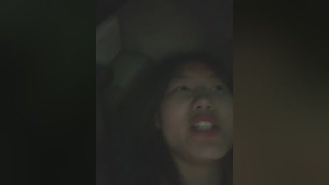 A blurry video of a young Asian woman with light skin, wearing a black headscarf, standing against a dark background. She appears distressed, eyes wide open and mouth agape, suggesting urgency or fear.
