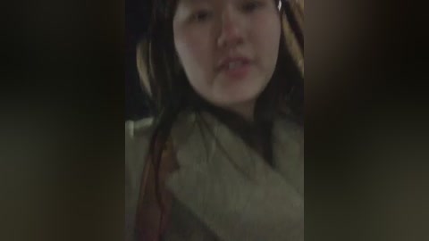 A dimly lit video of a young woman with fair skin, brown hair, and a serious expression. She wears a beige scarf and a muted, possibly historical, outfit. The background is dark and blurred, focusing attention on her face.