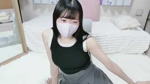Video of an East Asian woman with long black hair, wearing a black tank top, gray skirt, and white face mask, sitting on a white bed in a minimalist bedroom with white walls and a large mirror.