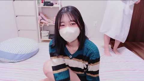 A video of an East Asian woman with long black hair, wearing a white mask, a striped sweater, and a blue dress, kneeling on a striped bed in a minimalist room with a white shelf and a wooden door.
