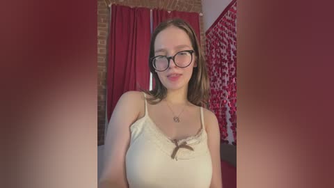 Video of a fair-skinned woman with glasses, light brown hair, wearing a beige camisole, standing in a room with red curtains, brick wall, and red floral wallpaper.