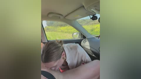 A candid video of a woman with blonde hair, wearing glasses, resting her head on the shoulder of a man with a visible tattoo. They are in a car with a green tint, driving through a lush, green landscape.
