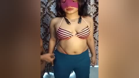Video of a young woman with medium skin tone and long dark hair, wearing a striped bikini top, blue leggings, and a pink mask, standing in a bathroom with floral-patterned shower curtain.