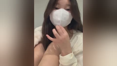Video of an Asian woman with long dark hair, wearing a white face mask, light-colored sweater, and beige pants, indoors against a blurred background.