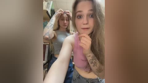 A candid photo of a young woman with long, wavy brown hair, wearing a pink tank top and high-waisted jeans, holding her chin. She has several tattoos on her arm. Background shows a cluttered room with books and a shelf.